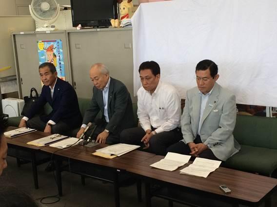 机の後ろの椅子に座って記者発表にのぞむ4人の男性の写真