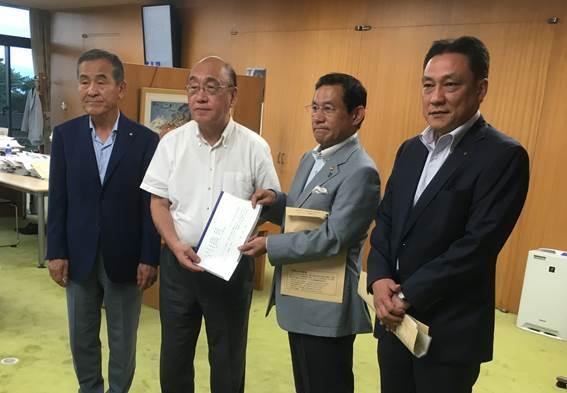 茶色い壁の部屋で真ん中の男性2人が決議書を持ち、その外にさらに2人の男性が一緒に写っている写真