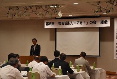 プロジェクターの前の演壇に立って講演するスーツ姿の男性の写真