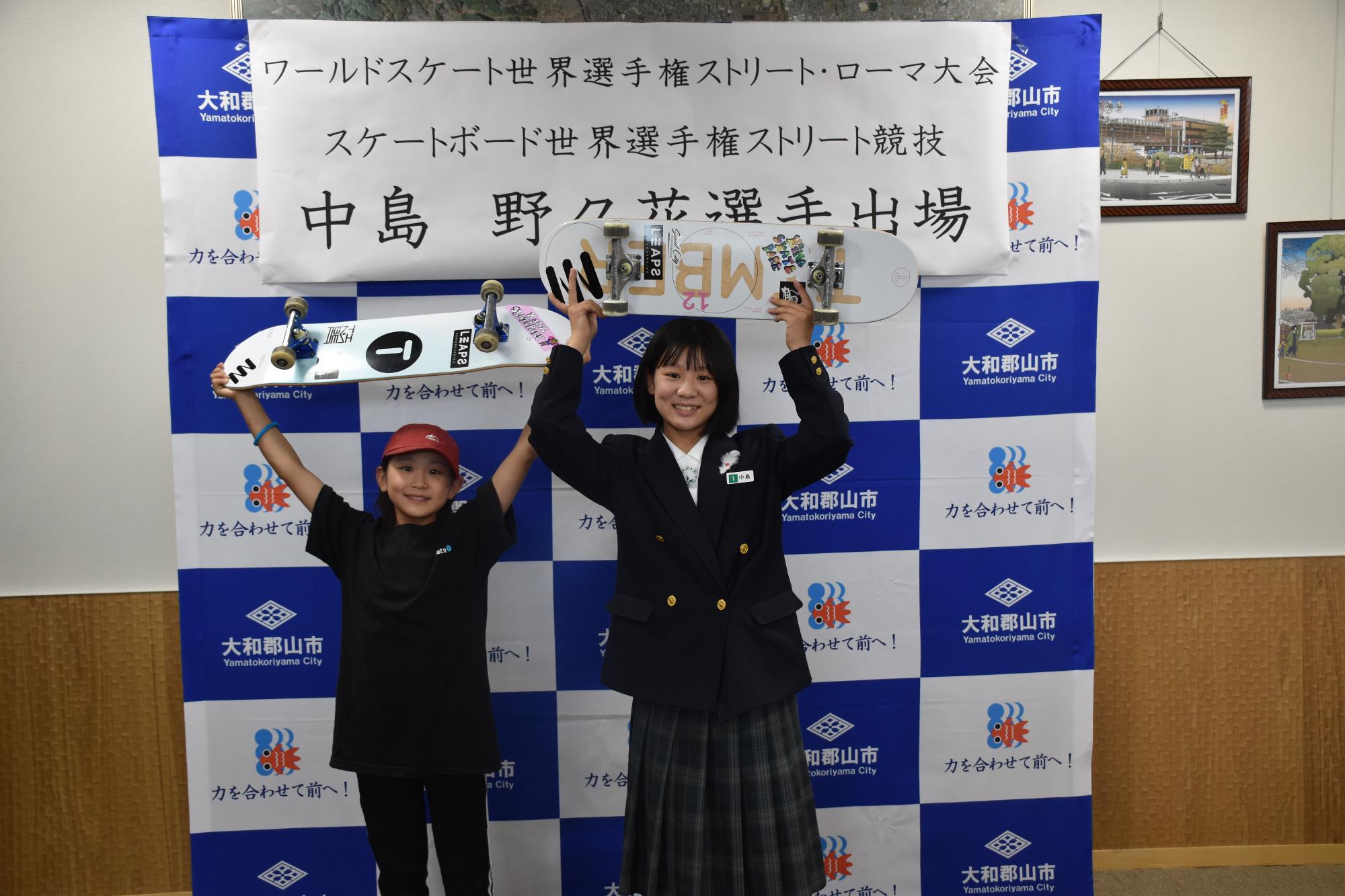 スケートボードを頭上に掲げて並んで立つブレザーの制服を着た野々花選手と赤い帽子と黒いTシャツの茉瑚選手