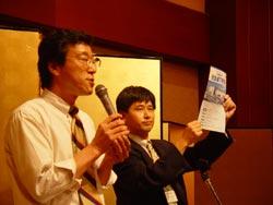 スピーチを行っている男性と元気城下町債を掲げている男性の写真