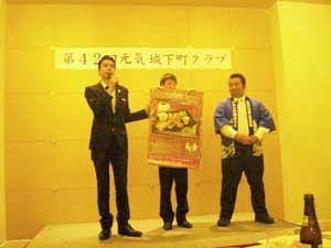 スーツの男性と法被姿の男性がポスターを持った男性を間でスピーチを行っている様子の写真