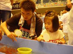 金魚すくい大会に参加しているお父さんの横で応援している女の子の写真