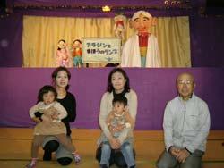 人形劇の登場人物をバックに記念撮影をする地域の2組の親子と児童委員の男性の写真