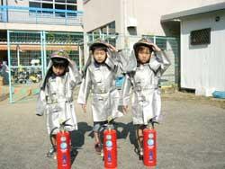 子供用の消防士の防護服をきて3本の消火器の後ろに立ち記念撮影をしている3人の園児たちの写真