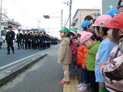 道路を整列して歩く消防団員を並んでみている子供たちの写真