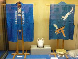 狐がプリントされた青いはっぴと狐のお面が展示されている写真