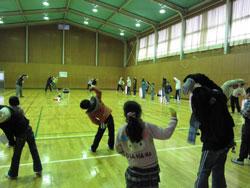 体育館でラジオ体操をする子どもたちが右脇の下を伸ばす姿勢をしている写真