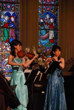 ステンドグラスの前でバイオリンとフルートを演奏する二人の女性の写真