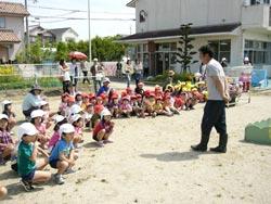 外で帽子をかぶったたくさんの子供たちがしゃがんで立っている男性を見ている写真。