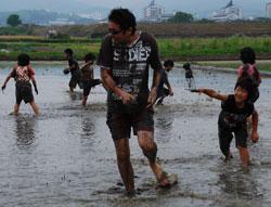 泥田の中を立っている男性に泥玉を投げている子供の写真