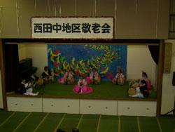 西田中地区敬老会と上に書かれたステージで、ピンクや白の衣装を着た人達が座って楽器を演奏している写真