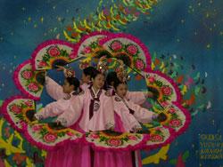 大きなイラストが飾られたステージで、ピンク色の衣装を着た人達が中央にあつまり、輪を作っている様子を大きく移した写真