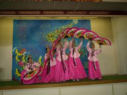 大きなイラストが飾られたステージで、ピンク色の衣装を着た人達が扇で波上の形を作っている写真