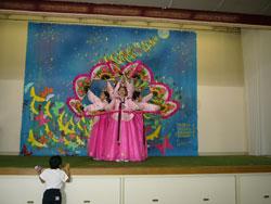 大きなイラストが飾られたステージで、ピンク色の衣装を着た人達が中央に集まり扇を輪上に掲げている写真