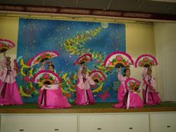 大きなイラストが飾られたステージで、ピンク色の衣装を着た人達が扇をもっている写真