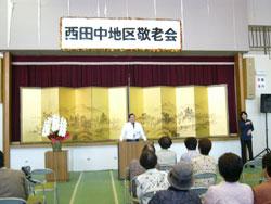 西田中地区敬老会と上に書かれたステージ前で、白い和服を着た男性が立っている、手前側にはその様子を座って見ている人達の写真