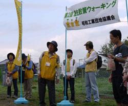 黄色い旗と白の横断幕の前にチョッキを着た人が複数名立っている写真