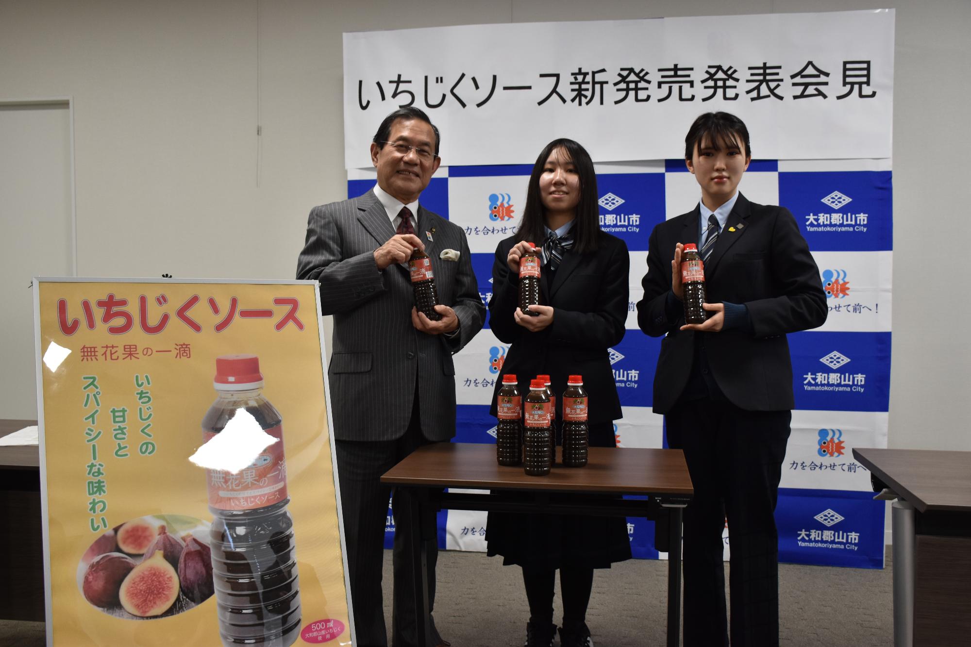 いちじくソースを手に持ち並ぶ上田市長と、紺色の制服を着た関西文化芸術高校の女子生徒2名