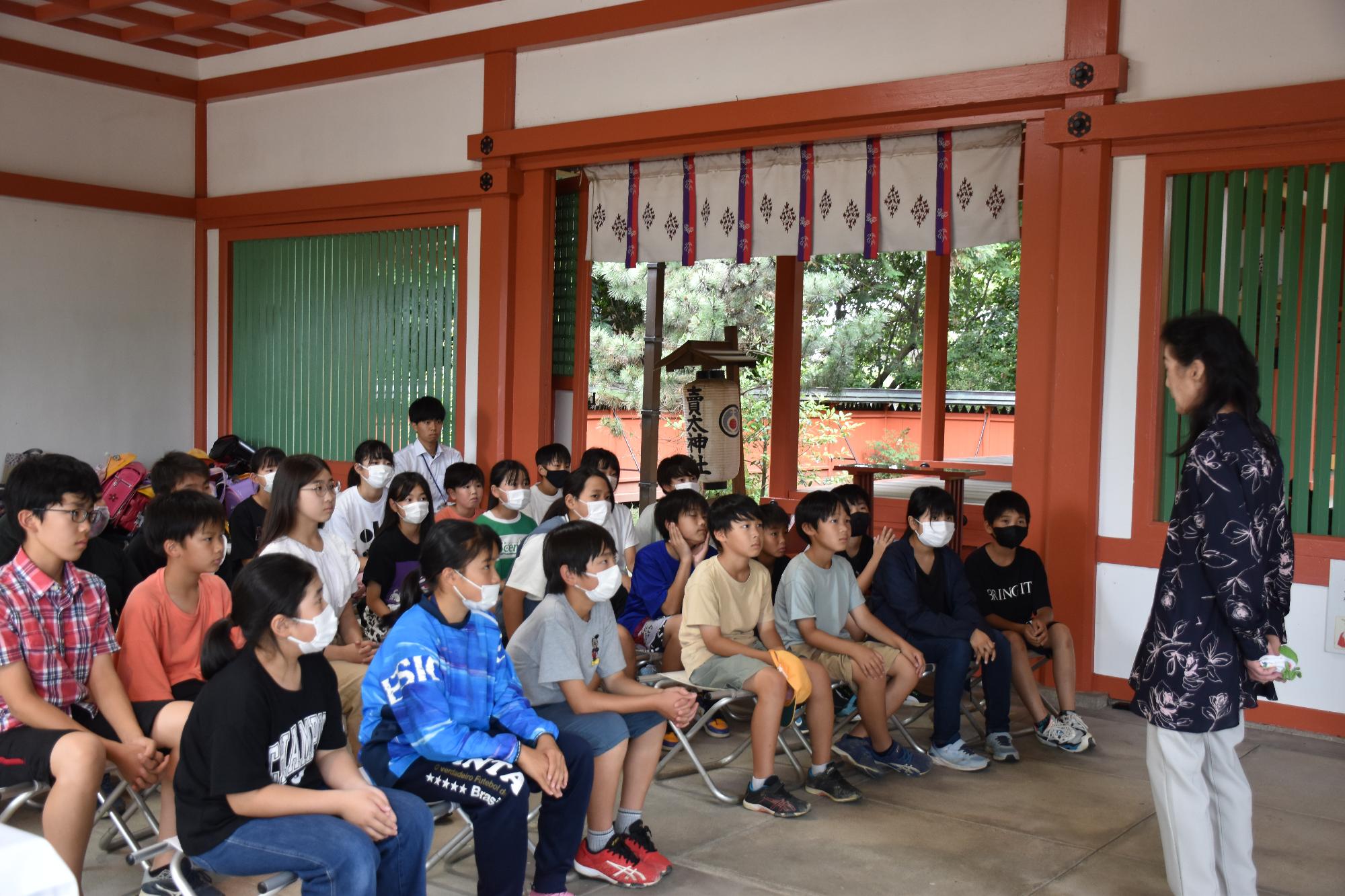 神社の本殿に並べられた椅子に座って、語り部の女性の話を聞く小学生児童たち