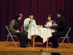 舞台でテーブルを囲んで演劇をする4人の人達の写真
