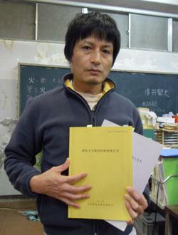 黒板がある部屋で黄色と白の2冊の冊子を持つ山川さんの写真