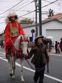 赤い衣服と金の甲冑を身に着けて、白い馬に乗る武田信玄役の男性の写真