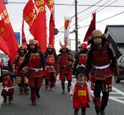六文銭の旗を背景に歩く、赤い甲冑を身に着けた男性と六文銭模様の服を着た子供の写真
