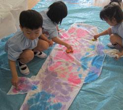 子供たちがこいのぼりに薄紫、ピンク、水色の絵の具を使って色を付けている写真