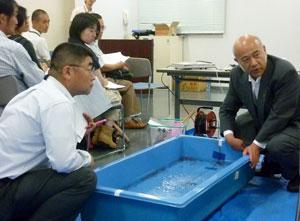 選手が水槽のどの位置にいればいいのかを実際に青い桶でレクチャーしている田村和勇氏のと審判員たちの写真