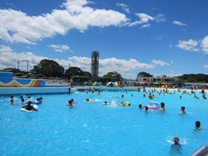 青空の下、たくさんの子どもたちが泳ぐ広いプールの写真