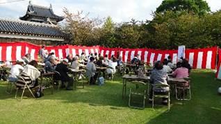 紅白の幕に囲まれ芝生の前庭にテーブルとパイプ椅子が並べられ参加者が着席している写真