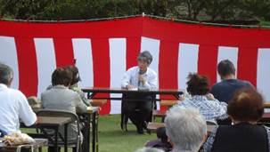紅白の幕のを背に参加者に向かい合い着席してマイクを持ち講演している男性の写真