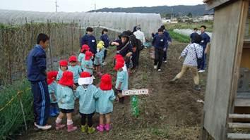 赤い帽子の複数の園児と青いジャージの中学生が畑の同じ場所で立っている写真