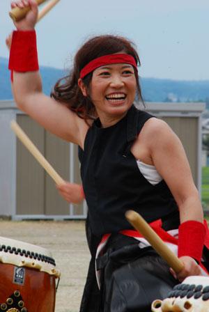 笑顔で太鼓を叩く赤い鉢巻をまいた女性の写真