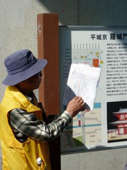 黄色いウェアを着た男性が平城京の羅城門跡を記す案内板の傍で説明書を広げている写真
