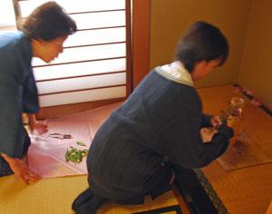 女子生徒が床の間に生花をさしており、後ろから年配の女性が見ている写真