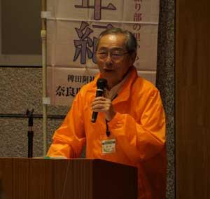 オレンジ色のジャケットを着た田原本ボランティアガイドクラブ会長がマイクを持って話している写真