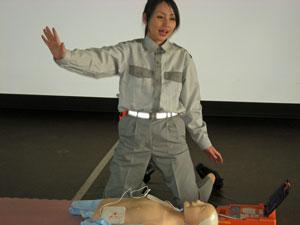 グレーの洋服を着た応急手普及員の女性が人形の前で両膝をつき片手を広げている写真
