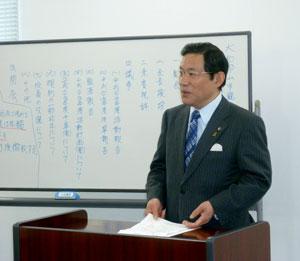 スーツを着た男性が、手に紙を持ち、ホワイトボードの前に立っている写真