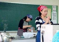 調理実習室で黒板の前にエプロンの女性、手前に赤い三角巾をつけた女性が立っている写真
