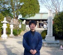 神社の前に青い作務衣で立つ宮司さんの写真