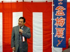 盆梅展の旗の横でスーツの男性がマイクを持って立っている写真