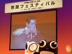 上田市長がハーモニカを吹いているプロジェクター映像とその前にいるゆるきゃらの写真