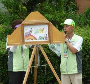 緑色のキャップの男性と、緑のチョッキを着た女性2人が紙芝居を披露している写真
