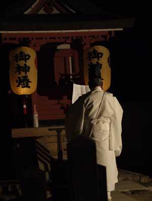 白い和服を着たご年配の男性が神を手もとで広げ、暗がりの神社で立っている写真