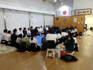 日本の国旗が飾られた広めの和室で輪になって会話をしている人たちの写真