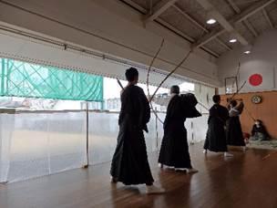 国旗が飾られた和室に袴を着た複数の人が弓を構えている、奥は屋外になっている場所の写真
