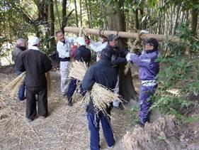 縄を編んでいる男性たちに材料の藁を渡そうとしている男性の写真