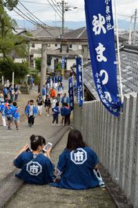 小泉神社秋祭りと書かれた青いのぼりがいくつもかかる柵がある坂道で、青い法被を着た2人の女の子が座り込んでいる様子を後ろから撮った写真
